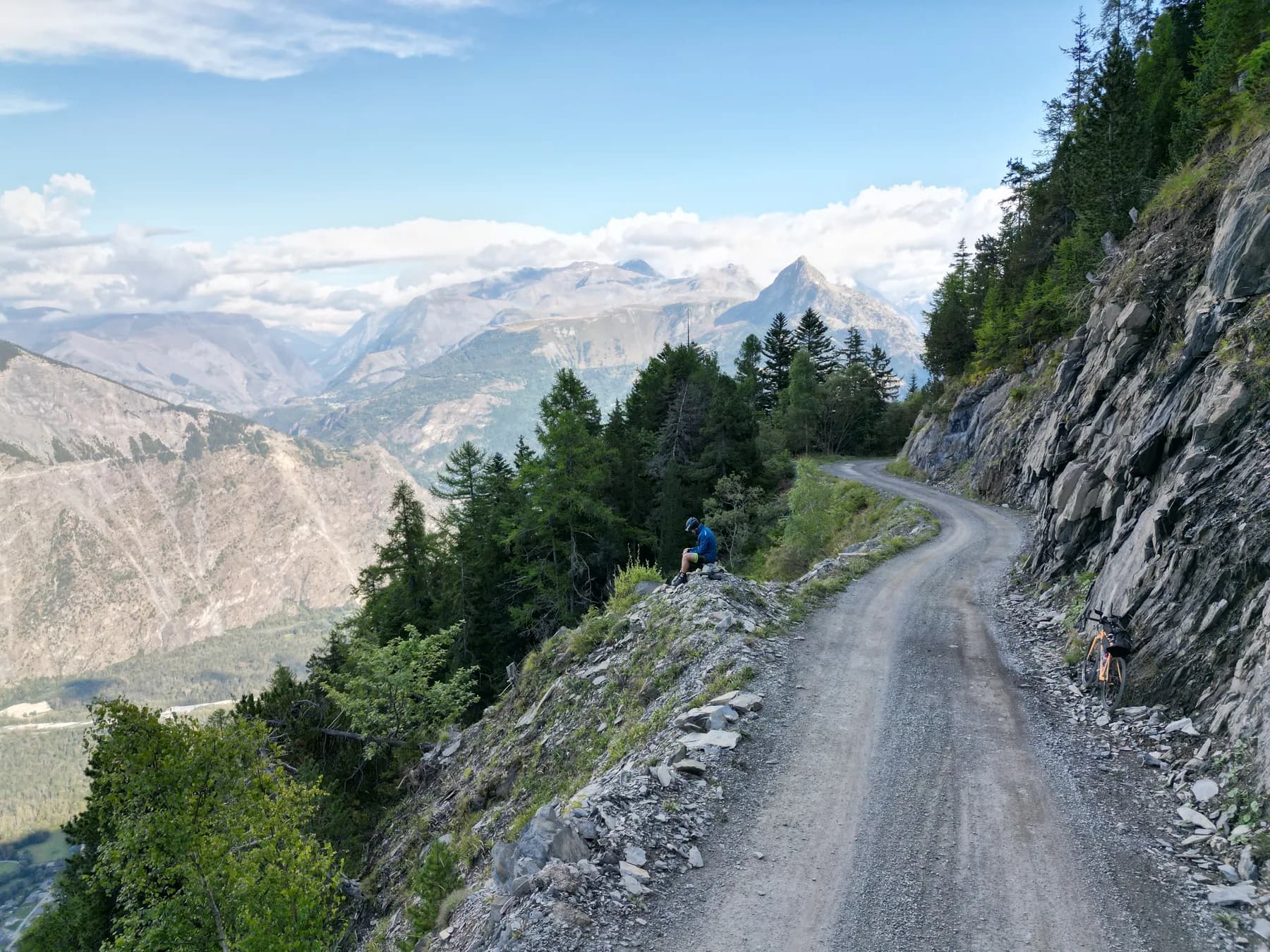 A cyclist in a blue jersey resting on a rocky outcrop beside a narrow gravel road carved along a steep alpine mountainside, a deep valley and distant peaks beyond and an orange gravel bike parked further along the track.