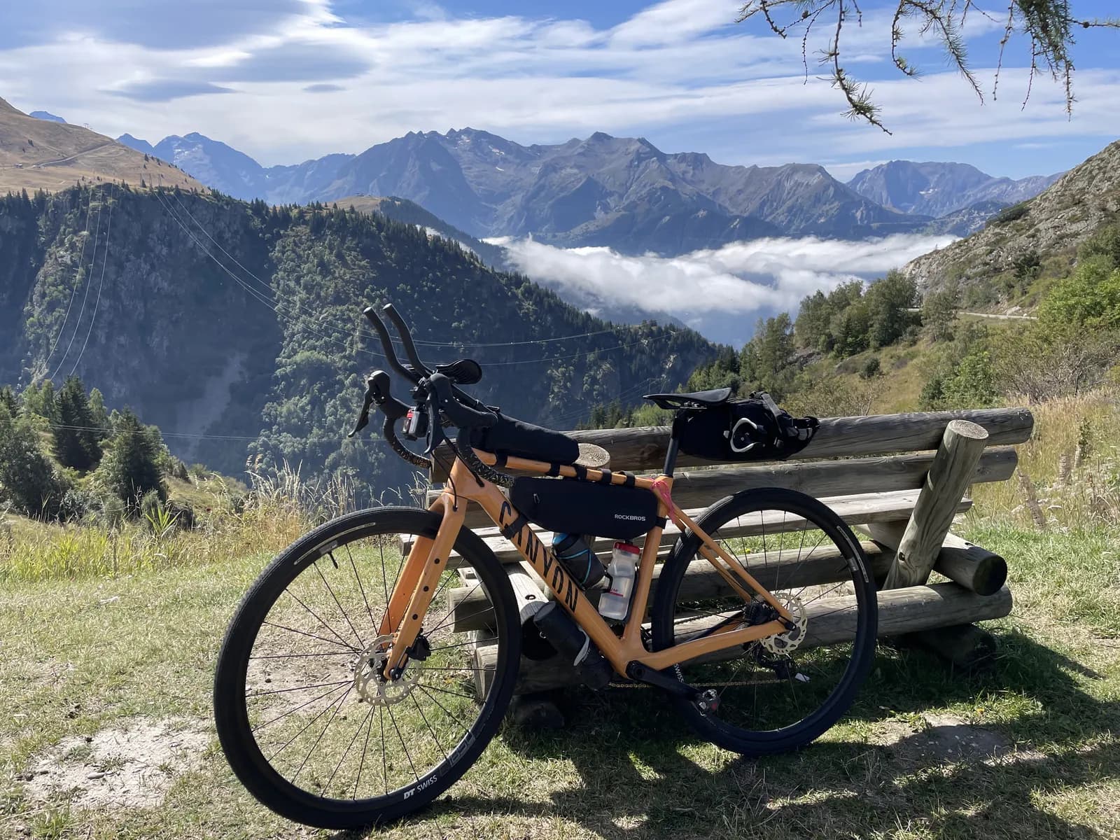 A loaded orange gravel bike leaning against a wooden log bench at an alpine pass, a cloud-filled valley below and snow-streaked peaks stretching to the horizon.